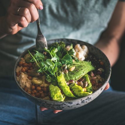 Healthy dinner or lunch. Woman in t-shirt and jeans eating vegan superbowl or Buddha bowl with hummus, vegetable, salad, beans, couscous and avocado, square crop